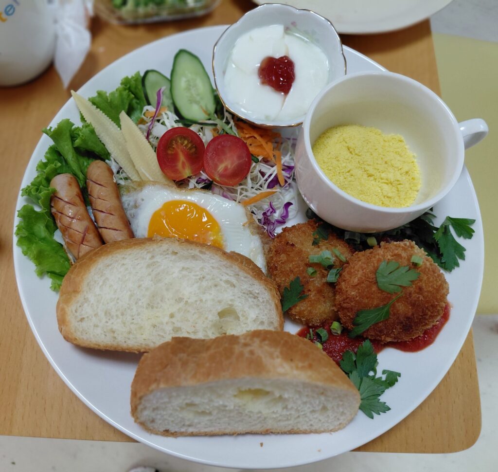 朝食ワンプレートに盛り付けられたパン、目玉焼き、ソーセージ、コロッケ、サラダ、ヨーグルト
