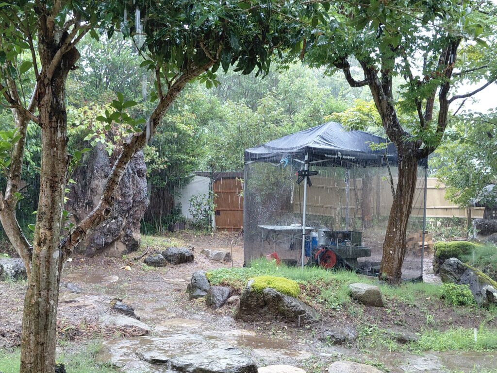 激しい雨に濡れる庭と、道具を守るタープテントの全景写真。